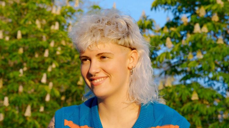 [ALT_TEXT: A young woman with a curly white blonde mullet haircut sitting outside on a sunny day.]