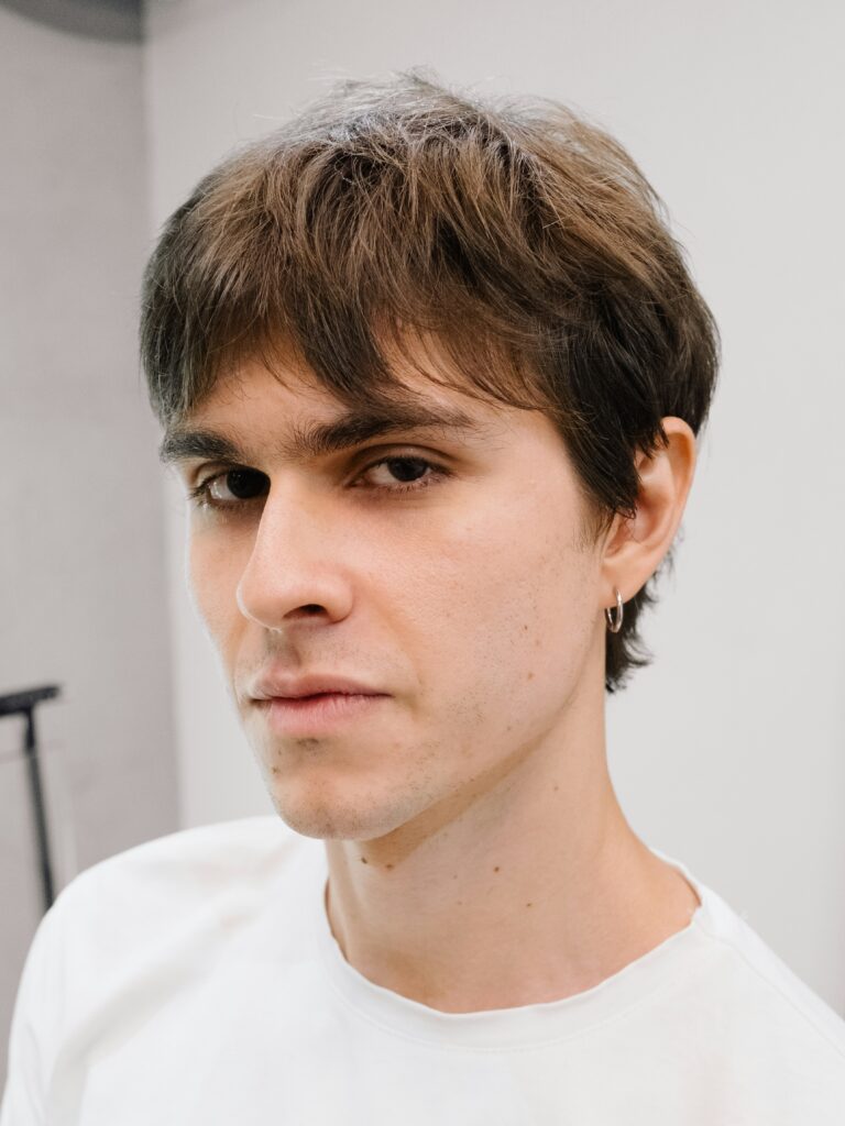 [ALT_TEXT: A young man with a business mullet haircut wearing a white t-shirt.]