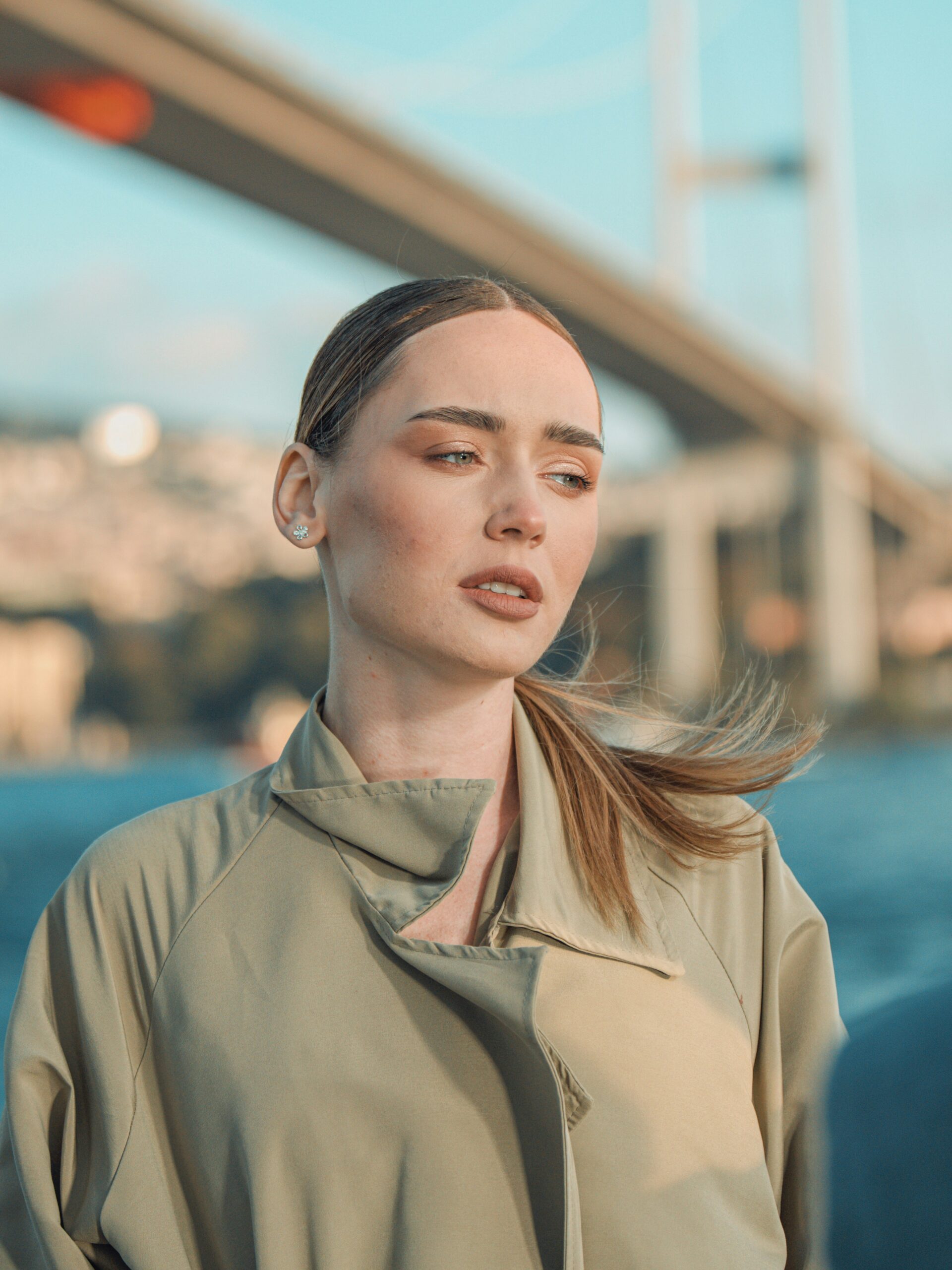 Μια γυναίκα με microblading στα φρύδια και ξανθά μαλλιά πιασμένα πίσω, φορώντας καπαρντίνα, μπροστά από μια γέφυρα στην Κωνσταντινούπολη