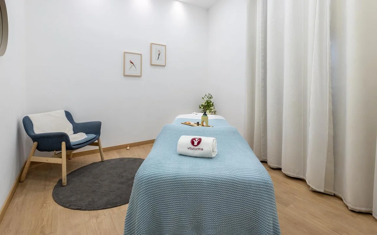 Ένα καθαρό, μινιμαλιστικό δωμάτιο spa με λευκούς τοίχους και ανοιχτόχρωμο ξύλινο πάτωμα. Ένα κρεβάτι θεραπείας είναι στρωμένο με μια γαλάζια πλεκτή κουβέρτα και μια τυλιγμένη λευκή πετσέτα με το λογότυπο «Vitaforma». Στη γωνία, μια κομψή μπλε πολυθρόνα πάνω σε μικρό στρογγυλό γκρι χαλί, ενώ μακριές λευκές κουρτίνες κρέμονται δίπλα στο παράθυρο.