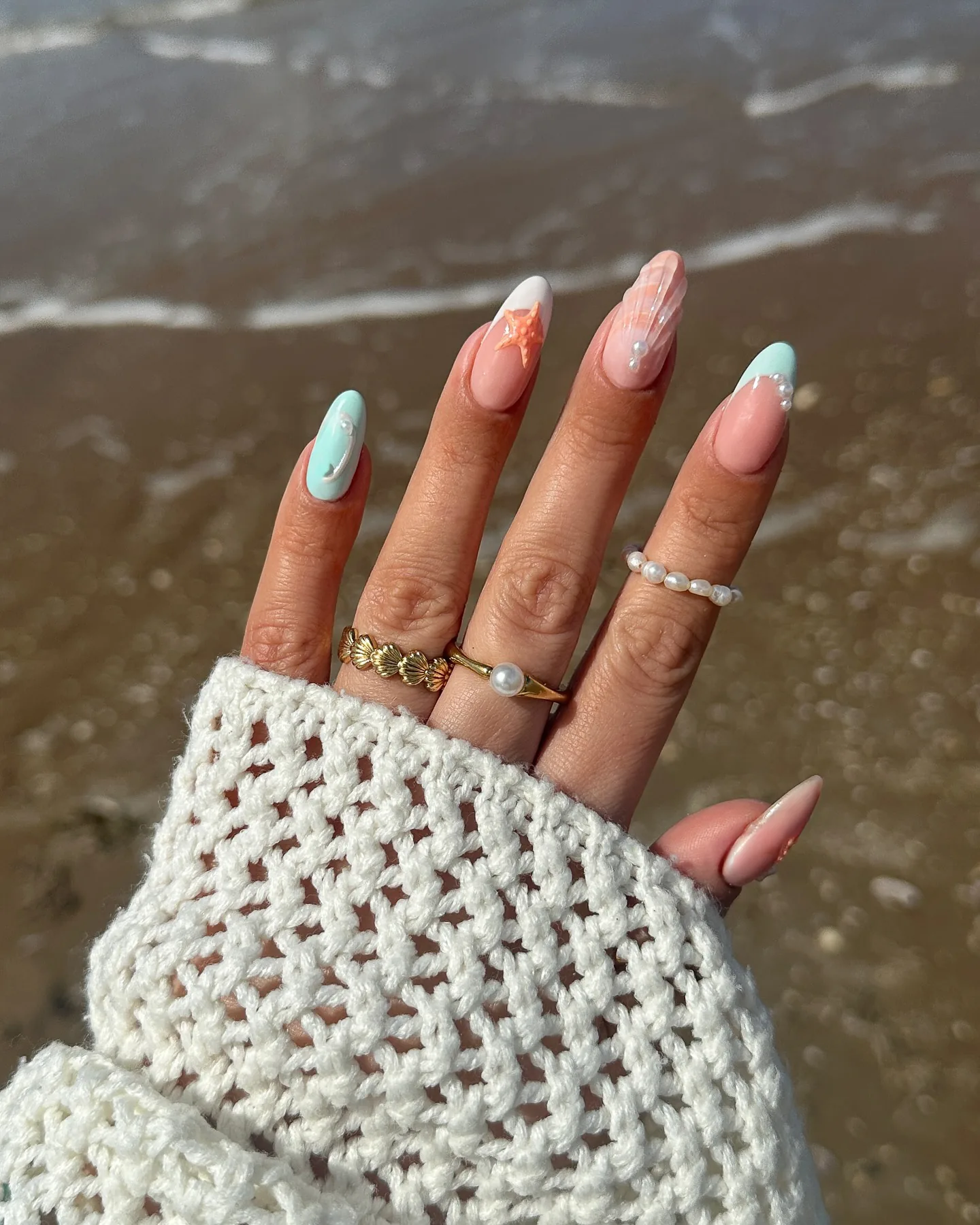 Αμυγδαλωτά νύχια με nail art γοργόνας σε παστέλ αποχρώσεις – έναν αστερία, περλέ πινελιές και ιριδίζουσα λάμψη – πάνω σε υγρή άμμο.