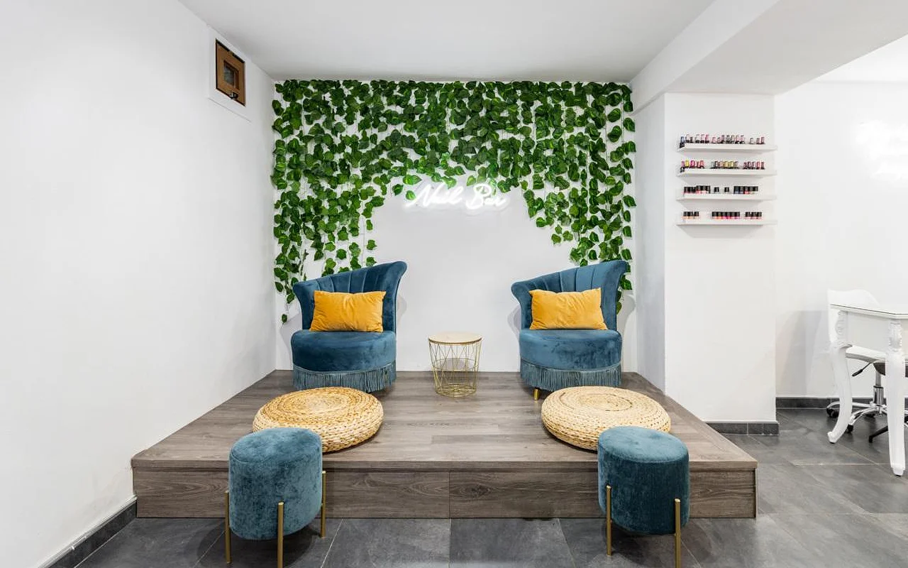 Μια γωνιά nail bar με έναν τοίχο από καταρρέον πράσινο φύλλωμα και μια μικρή πινακίδα νέον, δύο βελούδινες πολυθρόνες σε χρώμα πετρόλ με μουσταρδί μαξιλάρια πάνω σε μια υπερυψωμένη ξύλινη πλατφόρμα, πλεκτά υποπόδια-πουφ και ράφια με πολύχρωμα βερνίκια νυχιών στα δεξιά.