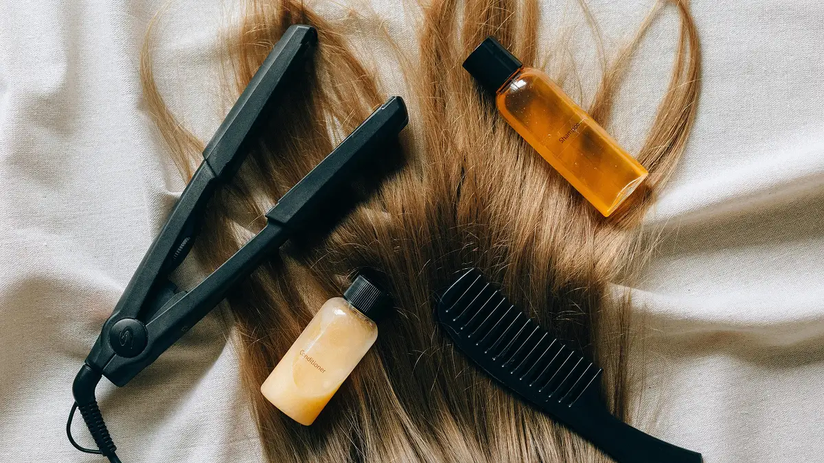 Flat lay προϊόντων περιποίησης μαλλιών τοποθετημένων πάνω σε μακριά ίσια μαλλιά, ανάμεσά τους σαμπουάν, conditioner, σίδερο και χτένα με αραιά δόντια