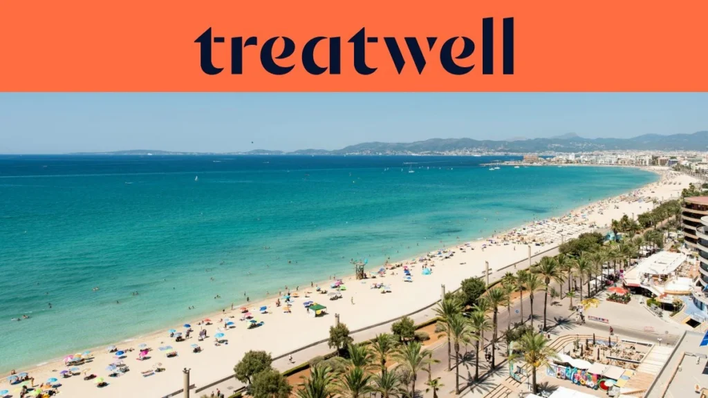 Αεροφωτογραφία της Playa de Palma στη Μαγιόρκα, με μια μακριά καμπύλη χρυσής άμμου, σειρές από ομπρέλες παραλίας, ήρεμα τιρκουάζ νερά και έναν παραλιακό περίπατο με φοίνικες κατά μήκος της πόλης, με λόφους στο βάθος κάτω από έναν καθαρό γαλάζιο ουρανό.
