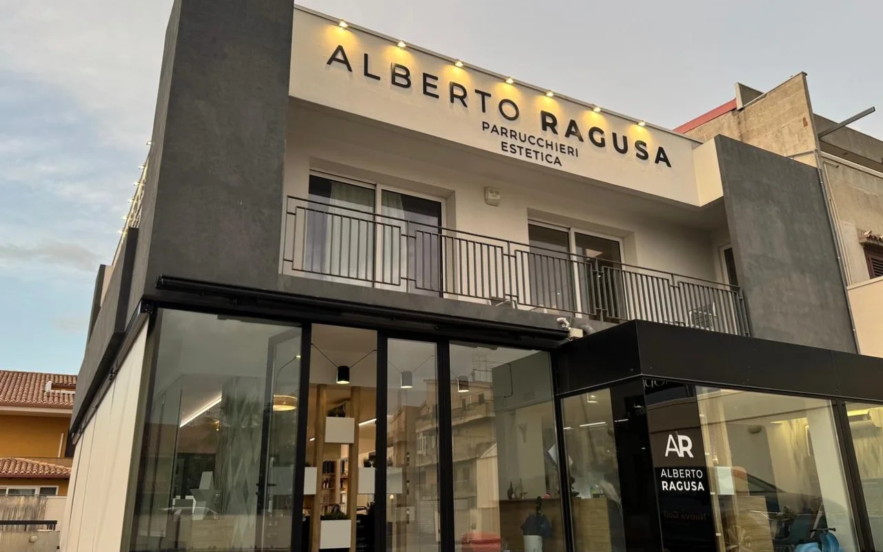 Η εξωτερική όψη του Alberto Ragusa το σούρουπο, ένα σαλόνι δύο ορόφων με μια μεγάλη φωτιζόμενη πινακίδα που γράφει «Alberto Ragusa Parrucchieri Estetica» πάνω από παράθυρα από το δάπεδο ως το ταβάνι, με ζεστό φως να λάμπει από μέσα.