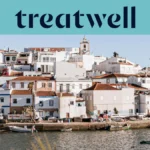 Ένα ασβεστωμένο ψαροχώρι του Algarve με σπίτια με κεραμοσκεπές που ανεβαίνουν σε μια πλαγιά πάνω από ένα ήρεμο νερό, μικρές ψαρόβαρκες δεμένες κατά μήκος της προβλήτας, φοίνικες και το καμπαναριό μιας εκκλησίας κάτω από έναν καθαρό γαλάζιο ουρανό.