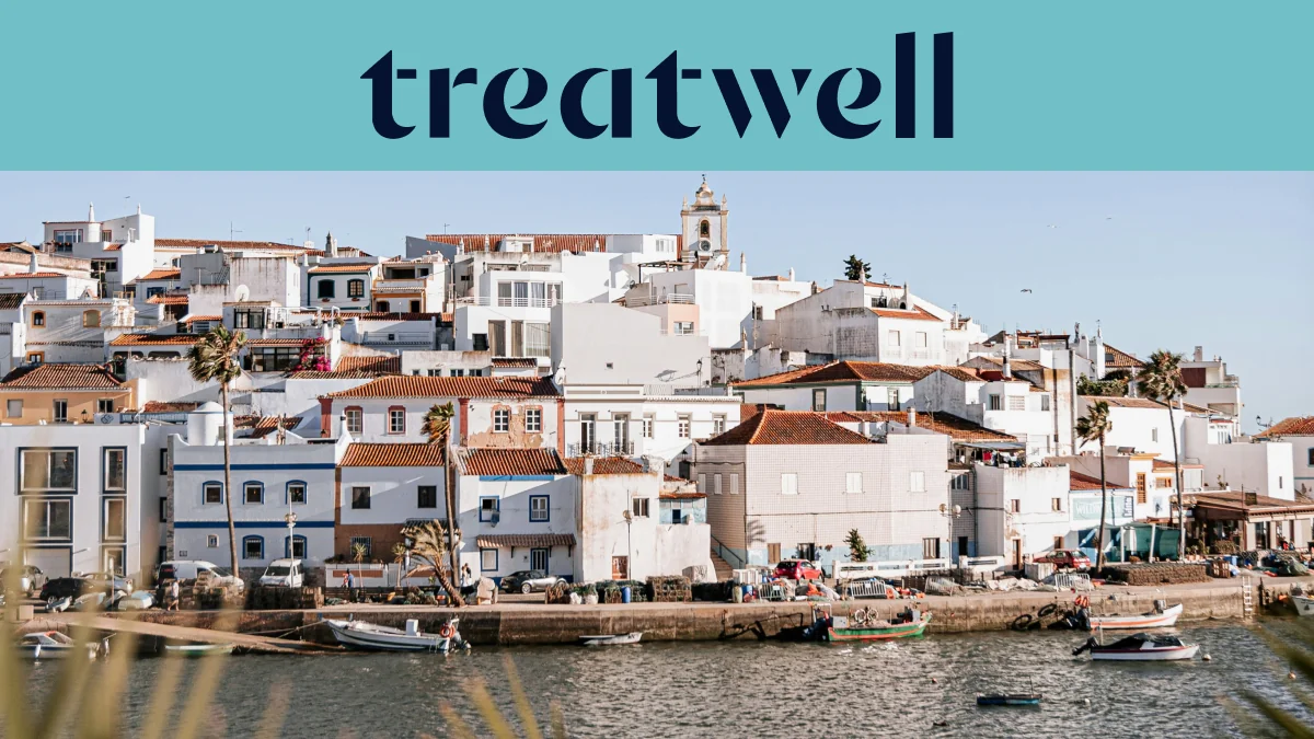 Ένα ασβεστωμένο ψαροχώρι του Algarve με σπίτια με κεραμοσκεπές που ανεβαίνουν σε μια πλαγιά πάνω από ένα ήρεμο νερό, μικρές ψαρόβαρκες δεμένες κατά μήκος της προβλήτας, φοίνικες και το καμπαναριό μιας εκκλησίας κάτω από έναν καθαρό γαλάζιο ουρανό.