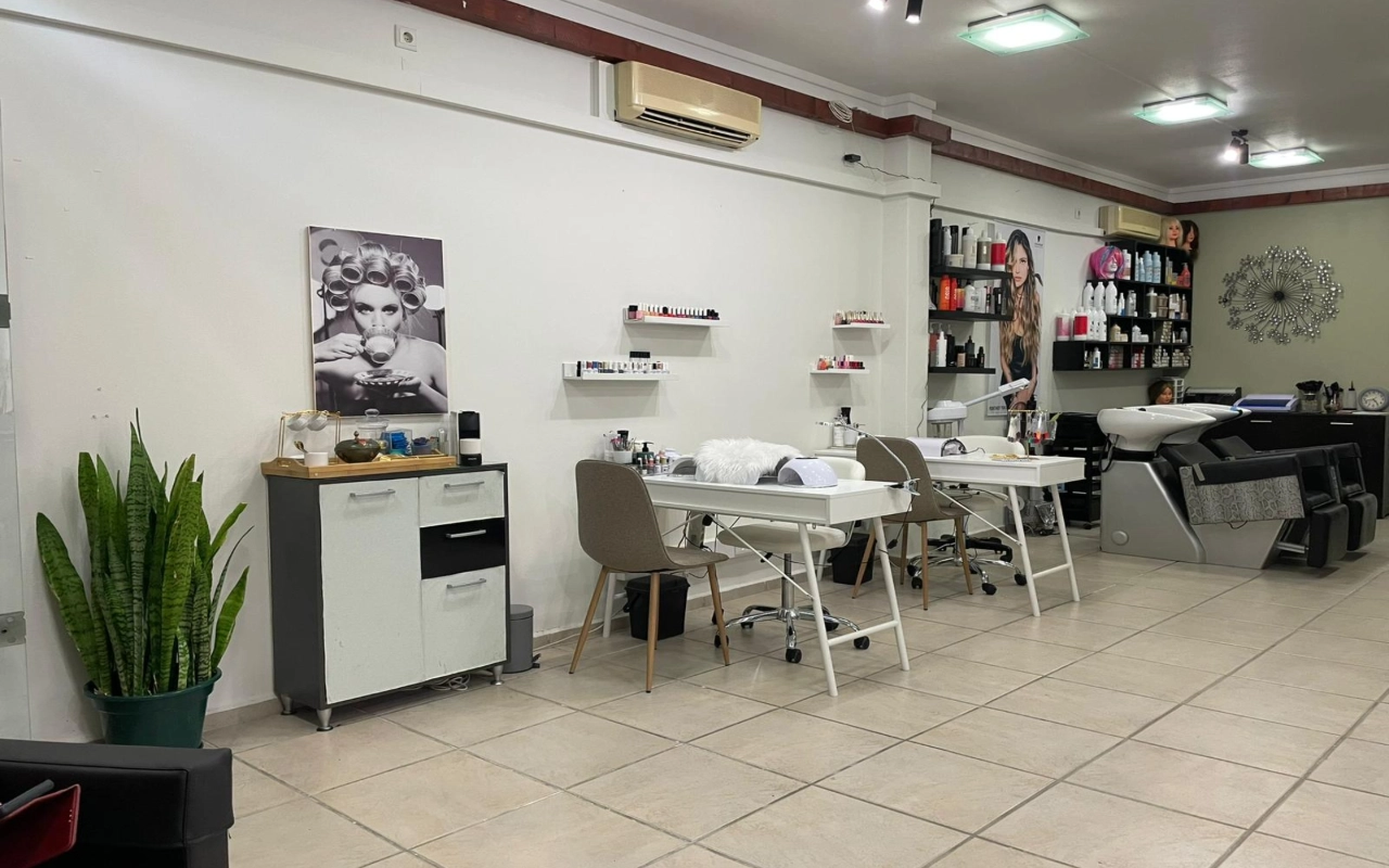 Ένα κομμωτήριο και σαλόνι ομορφιάς με δύο λευκά τραπέζια μανικιούρ και καρέκλες σε χρώμα τόπε, μια λεκάνη λουσίματος και ράφια προϊόντων κατά μήκος του τοίχου, ένα ψηλό φυτό σανσεβιέρια σε μια γωνιά και μια μεγάλη ασπρόμαυρη φωτογραφία μιας γυναίκας με ρόλεϊ.