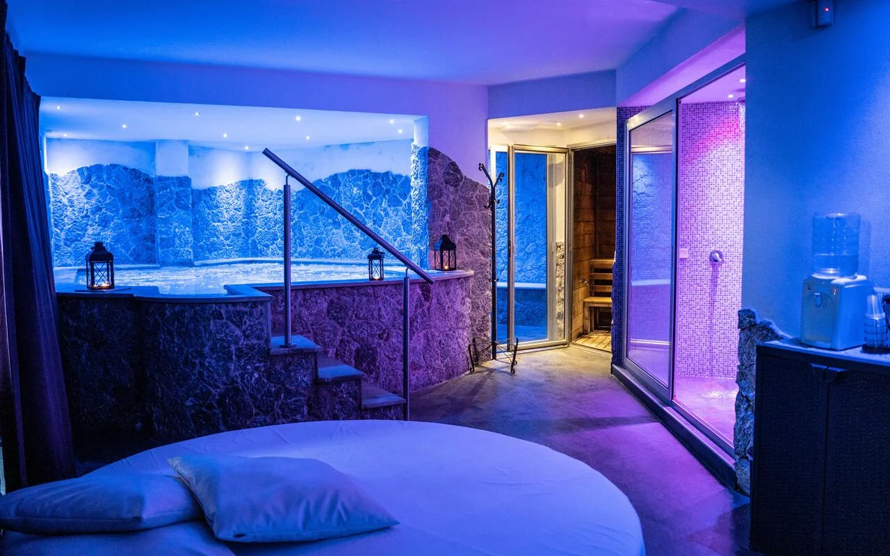 Μια ιδιωτική σουίτα spa στο Bellezza e Benessere στην Ταορμίνα, φωτισμένη σε μπλε και μοβ, με ένα εντοιχισμένο υδρομασάζ σε πέτρινη επένδυση, μια ξύλινη σάουνα και πίσω της ένα χαμάμ με ψηφιδωτά πλακάκια, και ένα στρογγυλό κρεβάτι στο προσκήνιο.