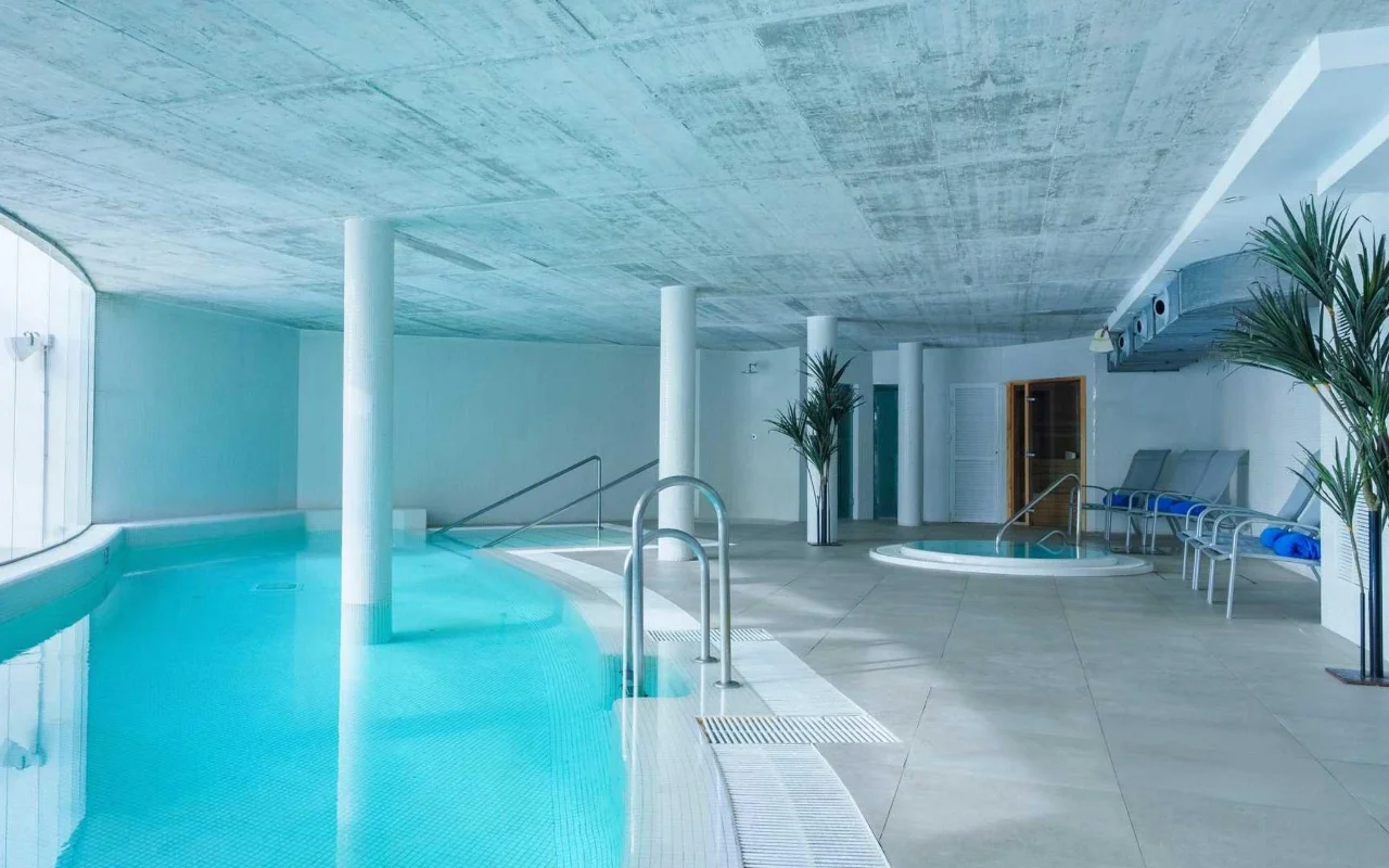 Η εσωτερική πισίνα spa της Benalmádena Beach, με μια καμπύλη τιρκουάζ πισίνα, ταβάνι από εμφανές μπετόν, ένα μικρό τζακούζι και ξαπλώστρες ανάμεσα σε ψηλούς φοίνικες σε γλάστρες.