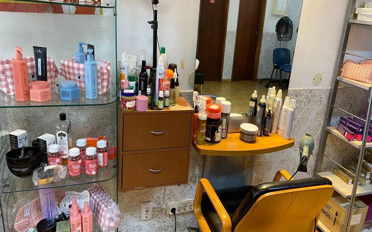 Ο σταθμός εργασίας ενός κομμωτή με έναν επιτοίχιο καθρέφτη και έναν ξύλινο πάγκο γεμάτο προϊόντα styling, μια γυάλινη βιτρίνα με προϊόντα περιποίησης μαλλιών και συμπληρώματα δίπλα, και μια κλασική καρέκλα κομμωτηρίου σε ξύλο και μαύρο.
