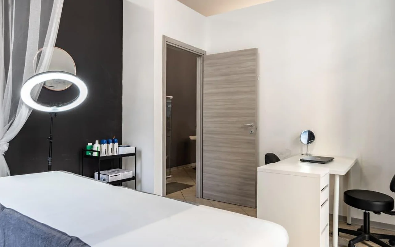 Μια καμπίνα θεραπείας στο Coquette Wellness and Beauty, με ένα λευκό κρεβάτι, ένα αναμμένο δαχτυλίδι φωτός σε τρίποδο μπροστά από έναν μαύρο τοίχο έμφασης, ελαφριές κουρτίνες στο παράθυρο και ένα μικρό λευκό γραφείο με καθρέφτη τουαλέτας δίπλα σε μια πόρτα που οδηγεί σε ιδιωτικό μπάνιο.