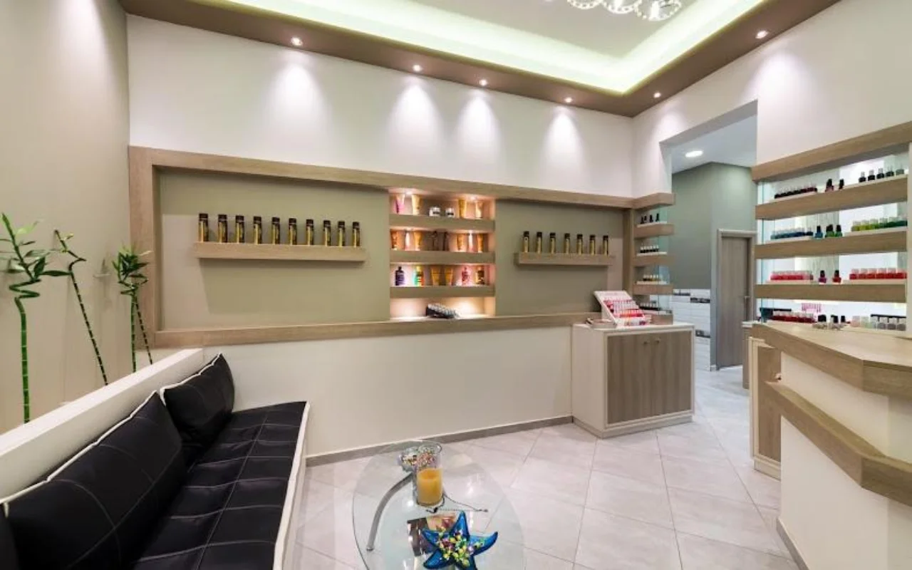 Η υποδοχή σε κρεμ αποχρώσεις στο Fabulous Nails and More στα Φηρά της Σαντορίνης, με έναν μαύρο δερμάτινο πάγκο, ένα γυάλινο τραπέζι, φωτιζόμενα ράφια με προϊόντα ομορφιάς και απαλό φωτισμό οροφής.