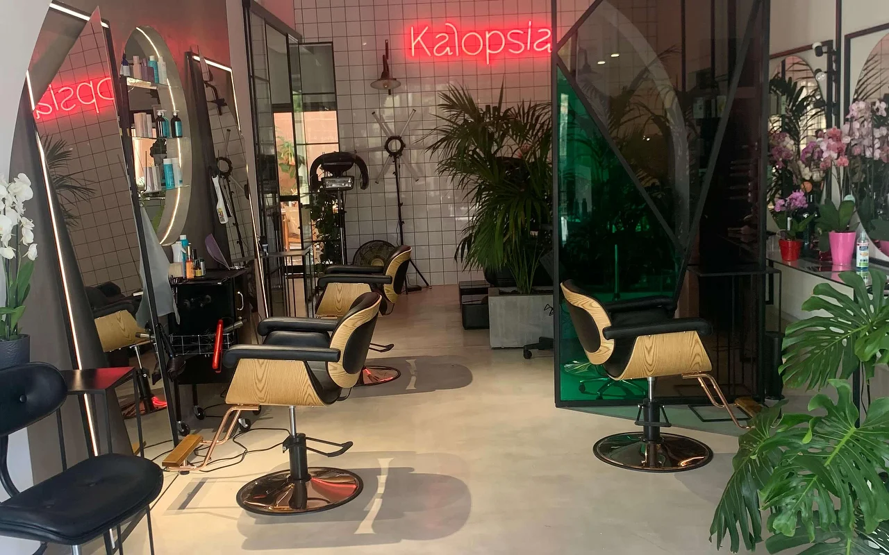 Ο χώρος styling στο κομμωτήριο Kalopsia στην Ερμούπολη της Σύρου, με καρέκλες σε ανοιχτό ξύλο και μαύρο, τοξωτούς φωτιζόμενους καθρέφτες, φυλλώδη φυτά και μια ροζ επιγραφή neon που γράφει Kalopsia.