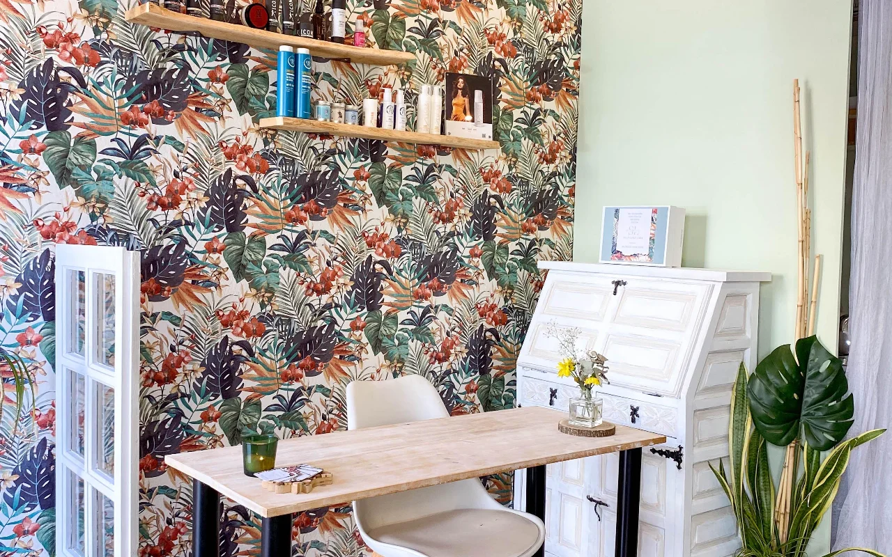 Μια θέση styling στη Shangara Lora Organics στη Marbella, με έντονη τροπική ταπετσαρία, ξύλινο τραπέζι, ένα παλιό λευκό γραφείο και βιολογικά προϊόντα μαλλιών σε αιωρούμενα ράφια.