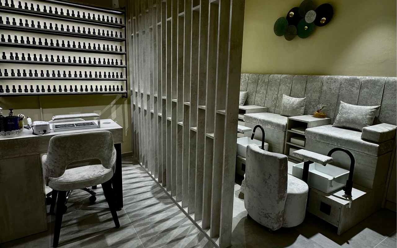 Ένα δωμάτιο περιποίησης στο Sisu Nail Club and Academy στην Περίσσα της Σαντορίνης, με έναν ολόκληρο τοίχο από βερνίκια νυχιών, γκρι βελούδινες καρέκλες μανικιούρ και πεντικιούρ και ένα τσιμεντένιο διάτρητο διαχωριστικό.