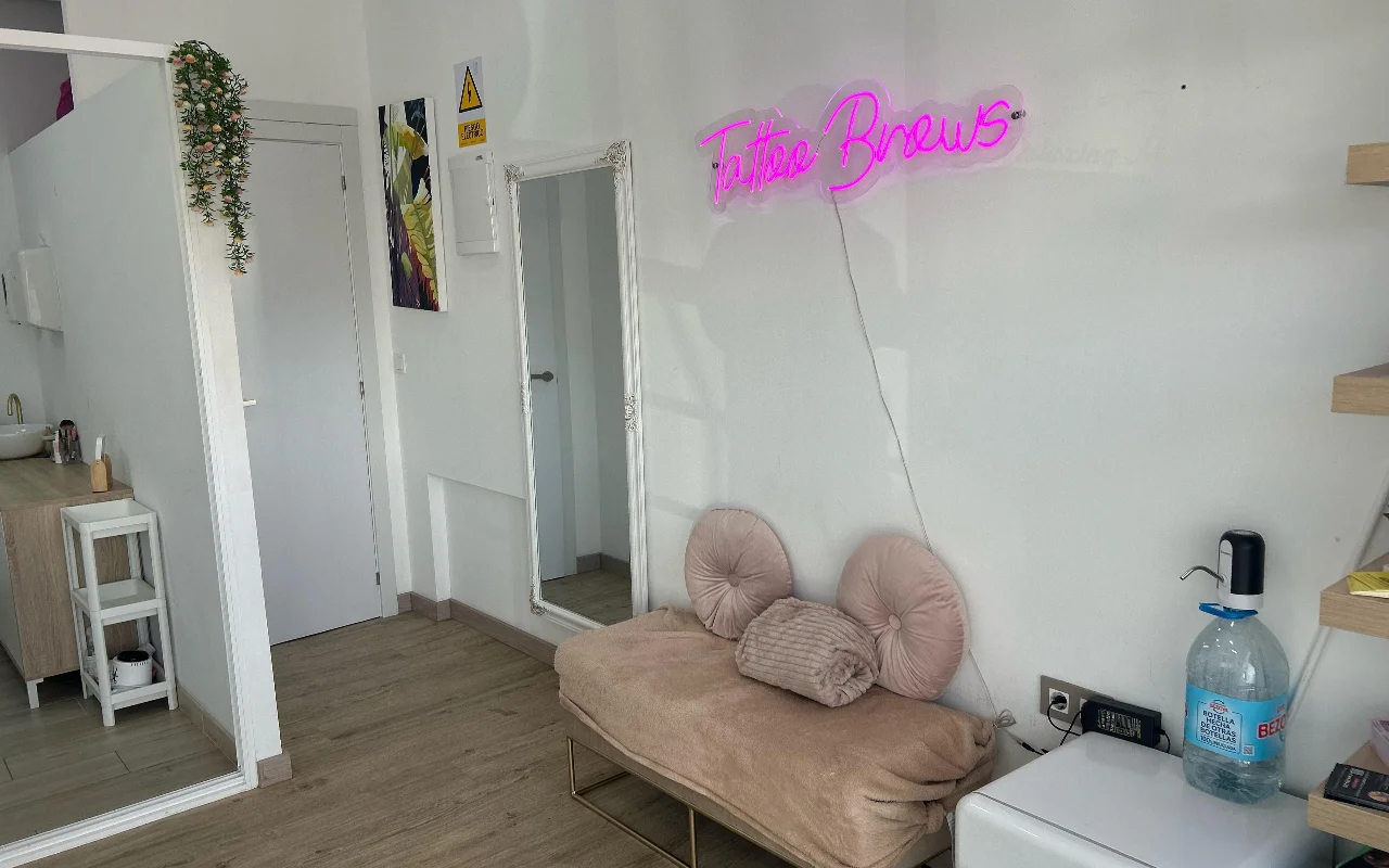 Ένας χώρος περιποίησης στη YG Beauty Estudio στη Torremolinos, με μια ροζ νέον επιγραφή «Tattoo Brows», έναν ολόσωμο καθρέφτη και μια μπεζ καπιτονέ πολυθρόνα.
