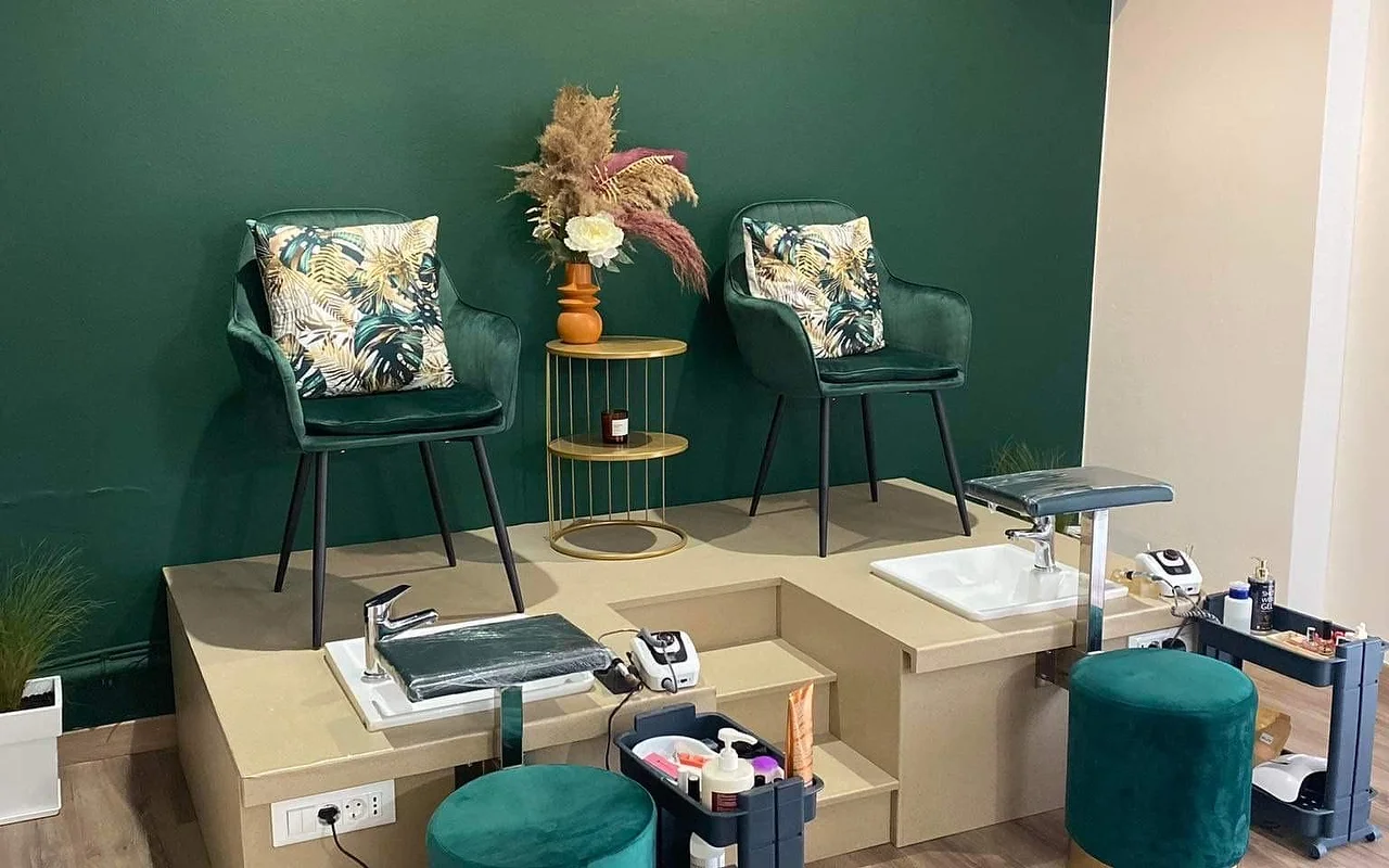 Μια γωνιά pedicure με βαθύ πράσινο τοίχο, δύο πράσινες βελούδινες πολυθρόνες κάδου με τροπικά εμπριμέ μαξιλάρια ανυψωμένες σε μπεζ πλατφόρμα πάνω από λεκανάκια pedicure, ένα χρυσό βοηθητικό τραπεζάκι που κρατά pampas σε πορτοκαλί βάζο, και ταιριαστά πράσινα βελούδινα σκαμπό.