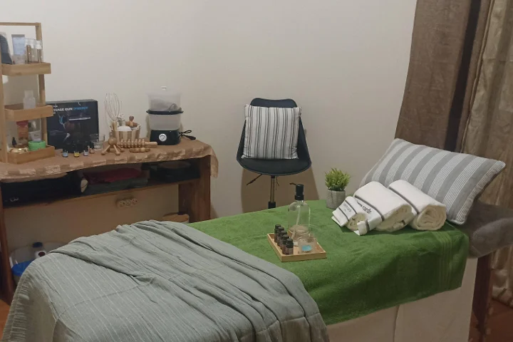 Ζεστός χώρος μασάζ με πολυθρόνα καλυμμένη με πράσινη πετσέτα και τυλιγμένες λευκές πετσέτες, δίσκο με αιθέρια έλαια, ξύλινα εργαλεία μασάζ σε ένα ρουστίκ βοηθητικό τραπέζι, και ένα σκαμπό δίπλα σε μια γωνιά χωρισμένη με κουρτίνα.