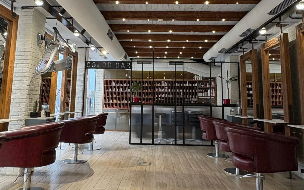 Ένα κομμωτήριο με εκτεθειμένα ξύλινα δοκάρια στην οροφή και ασβεστωμένο τούβλο, καρέκλες hair-styling σε δέρμα μπορντό, και ένας χώρος για βαφές με γυάλινους τοίχους καλυμμένος με ράφια προϊόντων πίσω από πινακίδα «Color Bar».